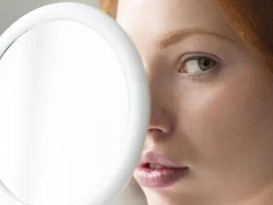 Девушка держит рядом с лицом зеркало, смотрит в кадр
